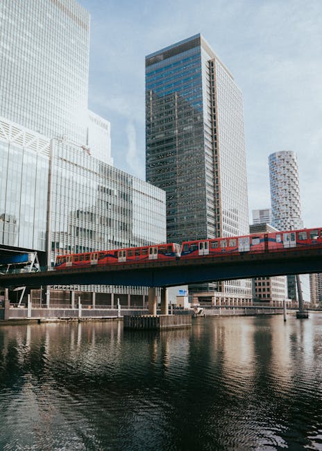 A modern cityscape featuring tall glass office buildings reflecting sunlight, with a red train traveling on an elevated railway bridge over a calm river. The buildings have smooth, shiny surfaces with