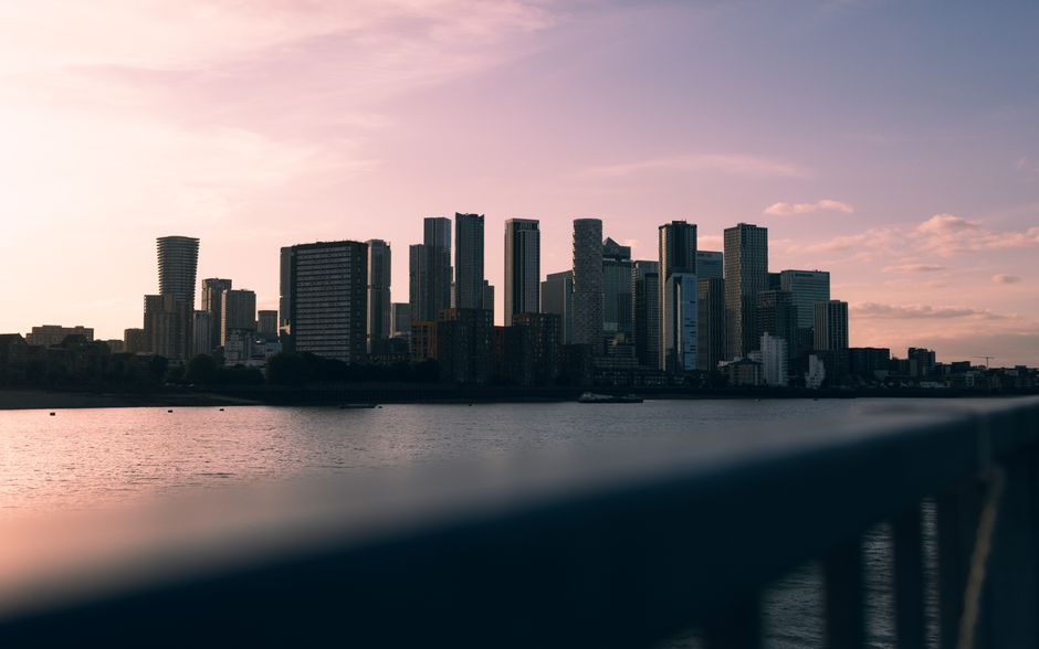 A modern cityscape featuring tall glass office buildings reflecting sunlight, with a red train traveling on an elevated railway bridge over a calm river. The buildings have smooth, shiny surfaces with