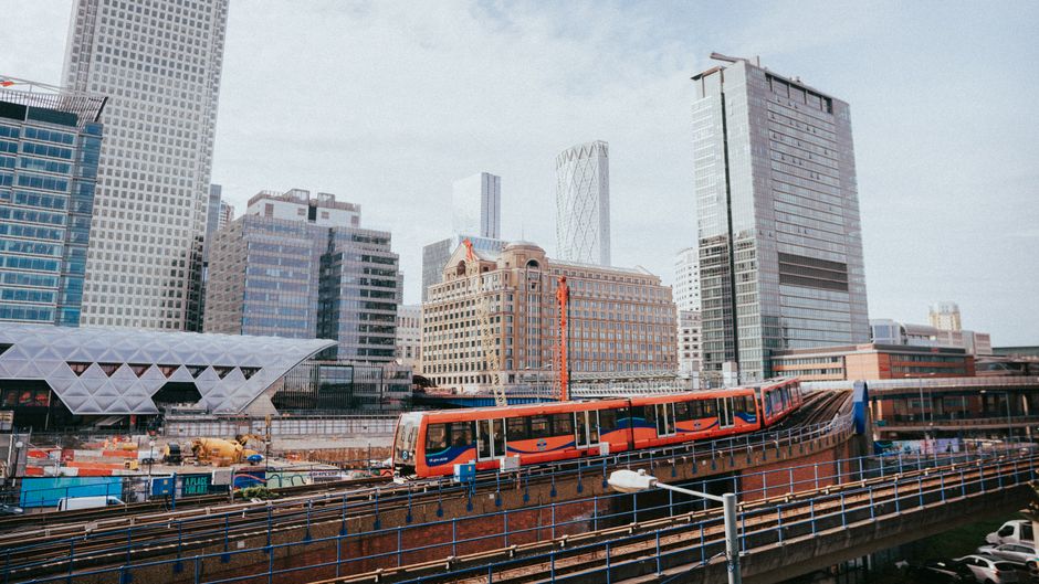 A modern urban scene in Canary Wharf featuring a sleek, orange and blue train on elevated tracks along with tall glass office buildings and contemporary architectural structures. The buildings include a mix of high-rise structures with reflective glass facades, one of which has a geometric white exterior, and another with a partially exposed steel framework at the top. The sky is partly cloudy with natural daylight illuminating the scene. The image emphasizes the city's modern infrastructure and the transport system, visible with metal railings and construction elements in the foreground. Canary Wharf Cleaners offers professional cleaning services near One Canada Square, ensuring the maintenance of such commercial environments.