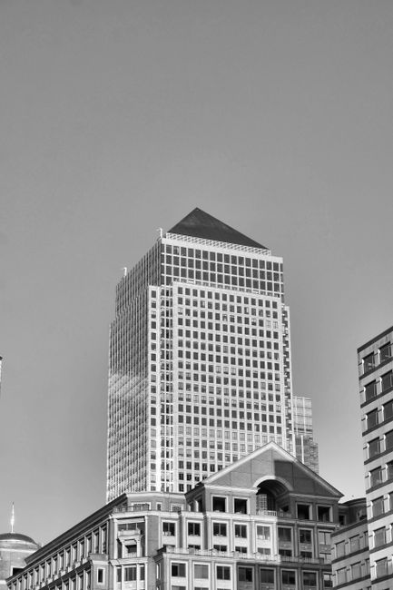 A grayscale photograph of a modern high-rise building located near One Canada Square in Canary Wharf. The building features a geometric façade with numerous rectangular windows arranged in a grid pattern, comprising glass and steel materials that reflect light. In the foreground, lower-rise structures with classic architectural elements, such as arched windows and decorative cornices, are visible. The scene is illuminated with natural daylight, highlighting the clean, well-maintained surfaces of the buildings. This image showcases urban architectural details relevant to Canary Wharf cleaning services offered by Canary Wharf Cleaners, emphasizing the importance of surface cleaning and maintenance in commercial environments.