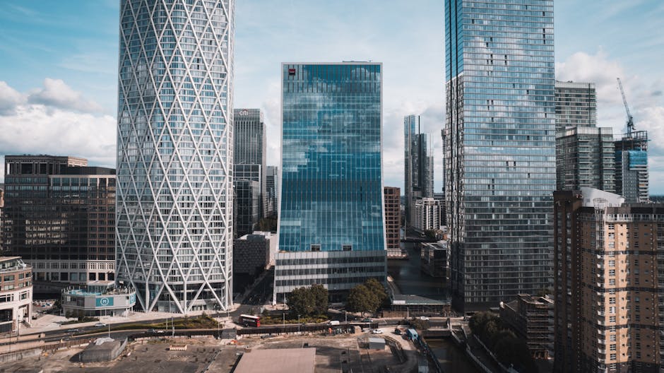 A modern urban scene in Canary Wharf featuring a sleek, orange and blue train on elevated tracks along with tall glass office buildings and contemporary architectural structures. The buildings include a mix of high-rise structures with reflective glass facades, one of which has a geometric white exterior, and another with a partially exposed steel framework at the top. The sky is partly cloudy with natural daylight illuminating the scene. The image emphasizes the city's modern infrastructure and the transport system, visible with metal railings and construction elements in the foreground. Canary Wharf Cleaners offers professional cleaning services near One Canada Square, ensuring the maintenance of such commercial environments.