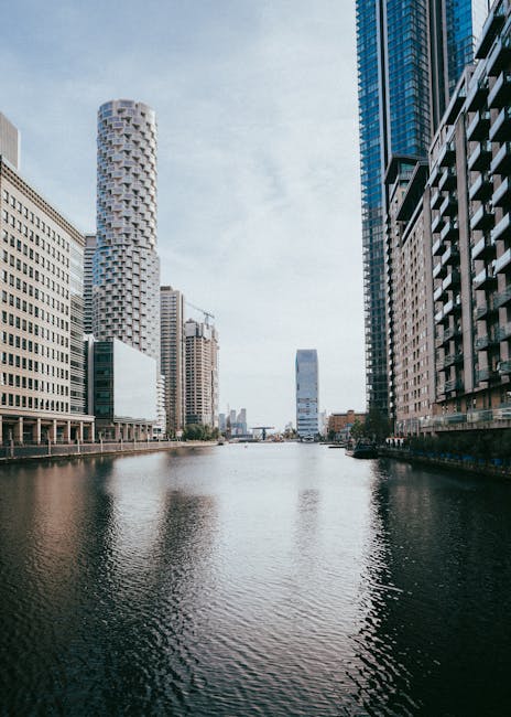 A wide view of the Heron Quays area in Isle of Dogs with tall modern office buildings on both sides of a calm River Thames. The buildings feature glass and steel facades, some with reflective windows, and vary in height and architectural style. The river surface appears clean and smooth, with gentle ripples, reflecting the sky and urban skyline. Natural daylight illuminates the scene, highlighting the sleek, polished surfaces of the structures. Canary Wharf Cleaners offers expert domestic and commercial cleaning services, including surface cleaning and deep cleaning, to maintain hygiene and shine in such high-end commercial environments.