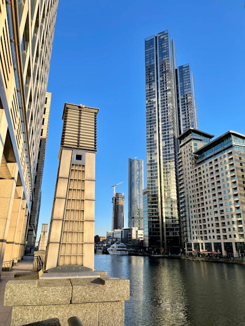 A view of tall modern skyscrapers along the water at Heron Quays, Isle of Dogs, featuring glass and steel facades reflecting sunlight. In the foreground, there are contemporary vent structures made of metal and concrete on a stone platform. The buildings vary in height, with some under construction, and the sky above is clear and blue. The image highlights an urban riverside scene with a focus on architectural details and clean, well-maintained surfaces, exemplifying professional surface cleaning and maintenance typical of Canary Wharf Cleaners in the commercial district.