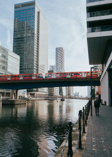 Image of a modern urban scene along the South Quay area of Canary Wharf, featuring a red London Underground train crossing an elevated railway bridge over the River Thames. The background showcases tall, reflective glass skyscrapers with sleek facades illuminated by natural daylight. On the right, a paved walkway runs parallel to the water, bordered by black metal posts connected by chains, with a person walking along the pathway. The water below the bridge is calm, reflecting the surrounding buildings and sky, creating a clean and organized cityscape. This scene emphasizes the contemporary architecture and busy transport infrastructure typical of Canary Wharf, aligning with the theme of surface cleaning, deep cleaning, and sanitisation associated with the South Quay end of tenancy cleaning guide. Canary Wharf Cleaners naturally provides expert cleaning services to maintain the hygiene and appearance of such commercial and residential environments.