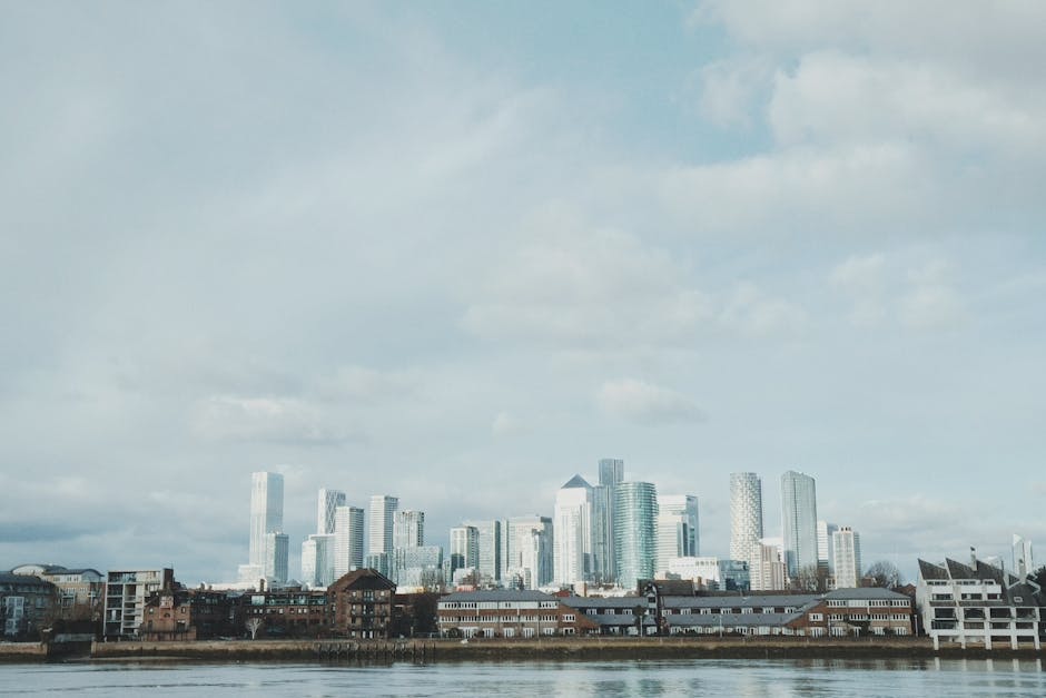 A cityscape view of Canary Wharf featuring modern skyscrapers with glass facades reflecting the cloudy sky, situated behind older brick and white buildings along the riverbank. The scene includes a mix of tall office towers and residential structures, with the water in the foreground showing calm reflections of the buildings. Bright natural light highlights the reflective surfaces of the commercial glass buildings, while the older structures add contrast. The image captures the architectural diversity and urban skyline typical of this area, with visible details of the building materials such as glass, brick, and steel. Canary Wharf Cleaners emphasizes surface cleaning and deep sanitisation, ensuring residential and commercial spaces remain pristine and hygienic, suitable for the vibrant business district shown in this image.