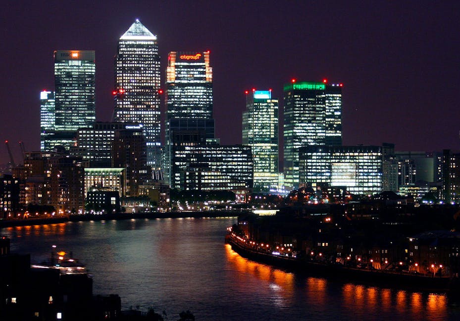 Image of a modern urban scene along the South Quay area of Canary Wharf, featuring a red London Underground train crossing an elevated railway bridge over the River Thames. The background showcases tall, reflective glass skyscrapers with sleek facades illuminated by natural daylight. On the right, a paved walkway runs parallel to the water, bordered by black metal posts connected by chains, with a person walking along the pathway. The water below the bridge is calm, reflecting the surrounding buildings and sky, creating a clean and organized cityscape. This scene emphasizes the contemporary architecture and busy transport infrastructure typical of Canary Wharf, aligning with the theme of surface cleaning, deep cleaning, and sanitisation associated with the South Quay end of tenancy cleaning guide. Canary Wharf Cleaners naturally provides expert cleaning services to maintain the hygiene and appearance of such commercial and residential environments.
